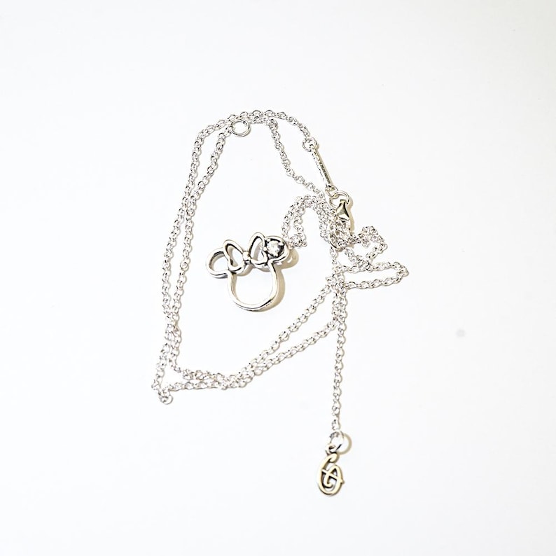 Puede incluir: Un collar plateado con un colgante con forma de cabeza y lazo de un personaje de dibujos animados. El collar tiene una cadena delicada y un peque&ntilde;o dije en el extremo. El colgante presenta una forma de cabeza circular con un lazo.