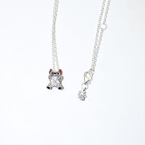 Op de afbeelding: Twee zilveren kettingen met een hanger met een stripfiguur. De hanger is een kleine Stitch-figuur van zirkonia met blauwe, rode en zilveren accenten. De ketting aan de rechterkant heeft een bloembedel en een sluiting.