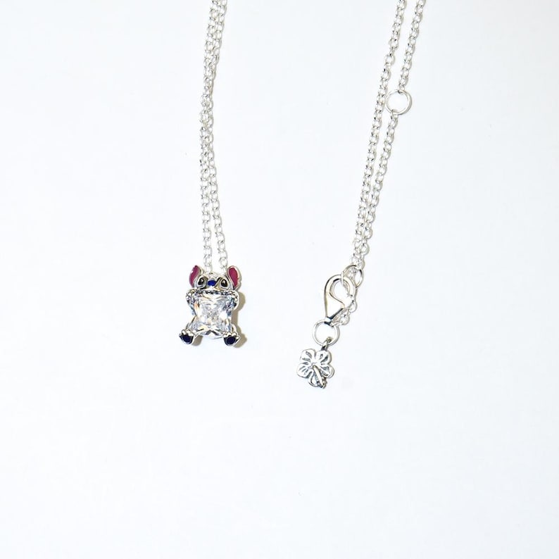 Op de afbeelding: Twee zilveren kettingen met een hanger met een stripfiguur. De hanger is een kleine Stitch-figuur van zirkonia met blauwe, rode en zilveren accenten. De ketting aan de rechterkant heeft een bloembedel en een sluiting.