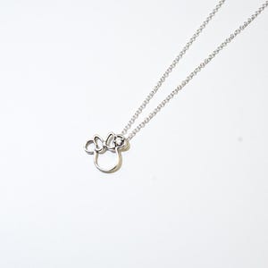 Puede incluir: Un collar plateado con un colgante de la cabeza de Minnie Mouse. El colgante es un contorno estilizado con un lazo y una peque&ntilde;a gema. El collar cuelga de una delicada cadena sobre un fondo blanco.