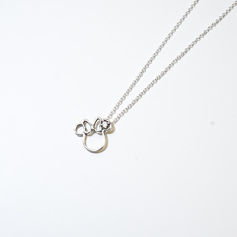 Puede incluir: Un collar plateado con un colgante de la cabeza de Minnie Mouse. El colgante es un contorno estilizado con un lazo y una peque&ntilde;a gema. El collar cuelga de una delicada cadena sobre un fondo blanco.