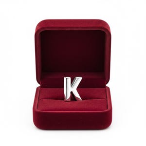 Può includere: Un anello argentato a forma di lettera "K" esposto in un portagioie di velluto bordeaux aperto. L'anello è centrato nella scatola, su sfondo bianco. Il portagioie è rettangolare con coperchio incernierato.