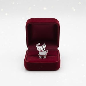 May include: A silver-colored robot-shaped charm is displayed inside an open, burgundy velvet jewelry box. The charm has a rectangular body, circular details, and a small, square chest. The box is set against a white background.