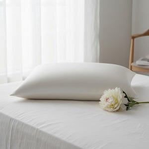 Puede incluir: Una almohada rectangular blanca descansa sobre una sábana blanca, con una flor blanca en primer plano. La almohada es lisa y parece estar hecha de un material suave. La escena está iluminada por luz natural, creando un ambiente sereno y tranquilo.