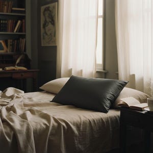Puede incluir: Escena de dormitorio con una cama con ropa de cama beige y una almohada gris oscuro. También se ve una almohada más clara. Un libro abierto y una taza están sobre una mesita de noche. Cortinas blancas transparentes enmarcan una ventana.