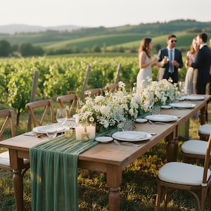 Peut inclure: Une longue table en bois dressée pour un événement extérieur, avec des assiettes blanches, des verres à vin et des centres de table floraux. Un chemin de table vert et des bougies ajoutent à l'ambiance élégante. Des invités sont visibles en arrière-plan, avec un vignoble au loin.