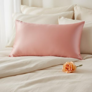 Puede incluir: Una funda de almohada de seda rosa rubor descansa sobre una cama con ropa de cama color crema. Una rosa color melocotón se encuentra cerca de la almohada. La funda de almohada tiene un acabado suave y brillante, lo que sugiere ropa de cama lujosa.