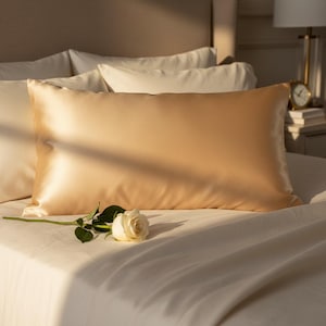 Puede incluir: Una almohada de seda dorada sobre una cama, con una rosa blanca y hojas verdes. La almohada tiene una textura suave y brillante. La ropa de cama es blanca.