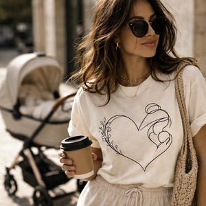 May include: Cream-colored t-shirt featuring a black line drawing of a heart with a mother and child silhouette and floral accents. The person is holding a brown coffee cup and wearing sunglasses. A stroller is in the background.