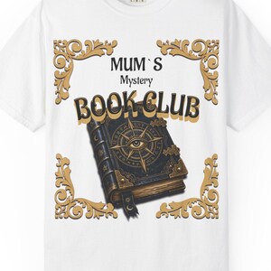 Könnte beinhalten: Ein weißes T-Shirt mit einem Grafikdruck eines dunklen, verzierten Buches mit Kompass- und Augenmotiv auf dem Einband. Der Text lautet MUM'S Mystery BOOK CLUB, umgeben von einer dekorativen goldfarbenen Filigranbordüre auf der Vorderseite.