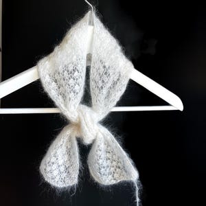Peut inclure: Une écharpe blanche et ajourée est présentée sur un cintre blanc sur fond noir. L'écharpe est nouée au centre, avec les extrémités pendantes. Une petite étiquette avec du texte est attachée à l'écharpe.