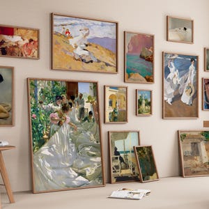 Op de afbeelding: Een verzameling ingelijste kunstdrukken in verschillende maten en stijlen, tentoongesteld op een beige muur. Het kunstwerk omvat landschaps- en figuratieve schilderijen met houten frames. Een kleine houten kruk en een tafel met een houten kom staan op de voorgrond.