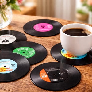 May include: A white mug filled with coffee sits on a black vinyl record coaster. Several other vinyl record coasters with colorful labels are scattered on a wooden surface. The coasters have various label designs.