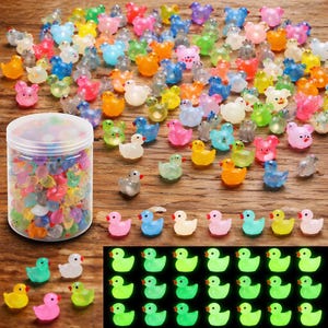 May include: A collection of colorful, miniature duck and bear figurines in various colors, including pink, blue, yellow, and green. Some are in a clear plastic container. A few glow in the dark. The figurines are scattered on a wooden surface.