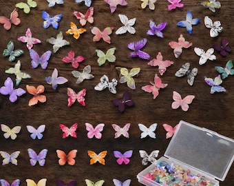 May include: A collection of colorful butterfly embellishments in various sizes and designs, scattered on a dark wood surface. The butterflies are in shades of pink, blue, yellow, and purple, with some stored in a clear plastic container.