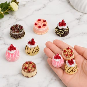 May include: A collection of miniature cake figurines in various designs, including chocolate, pink, and cream-colored options topped with red berries, white frosting, and drizzled icing, displayed on a white marble surface.