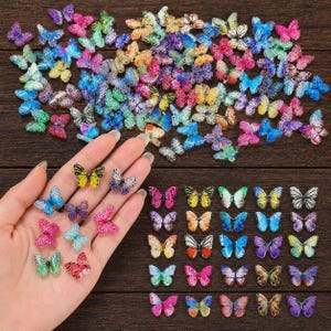 May include: A collection of colorful butterfly embellishments in various sizes and shades, including pink, blue, yellow, and green. Some butterflies are held in a hand, while others are scattered on a wooden surface. These decorative butterflies are ideal for crafts.
