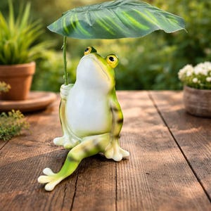 Puede incluir: Una caprichosa figura de rana, sosteniendo una gran hoja verde sobre su cabeza. La rana es blanca con detalles verdes y amarillos, y tiene grandes ojos amarillos. La figura está sobre una superficie de madera, con plantas al fondo.