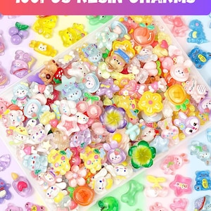 May include: A collection of colorful resin charms in various shapes, including flowers, animals, and fruits. The charms are in a clear plastic container, with the text "100PCS RESIN CHARMS" at the top. The charms are in shades of pink, yellow, blue, and green.