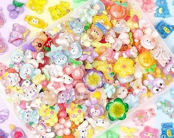 May include: A collection of colorful resin charms in various shapes, including flowers, animals, and fruits. The charms are in a clear plastic container, with the text "100PCS RESIN CHARMS" at the top. The charms are in shades of pink, yellow, blue, and green.