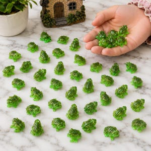 May include: A collection of small, bright green miniature frog figurines with textured skin, scattered across a surface, with several of the tiny plastic amphibians held in a palm to demonstrate their diminutive scale and detailed appearance.