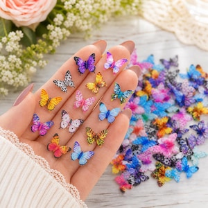 May include: A selection of colourful butterfly embellishments in various sizes and patterns. The butterflies are in shades of pink, blue, yellow, and purple, with some featuring speckled designs. Some butterflies are held in a hand, while others are scattered.
