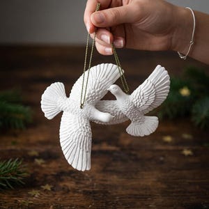 May include: Two white dove ornaments crafted with detailed feather textures on their wings and bodies. Each bird is suspended by a thin, metallic gold cord, allowing them to hang in a flying position with wings spread wide against a dark background.