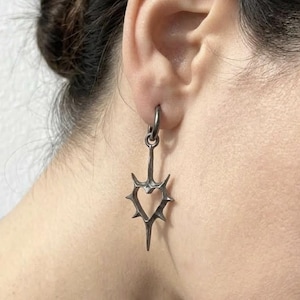 May include: A dark silver earring with a heart-shaped design, featuring thorn-like spikes around the heart's perimeter. The earring is suspended from a small hoop, creating a gothic or edgy aesthetic. The earring is worn on the ear.