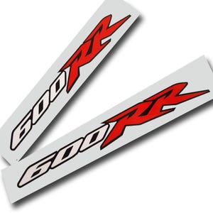 Könnte beinhalten: Zwei weiße Aufkleber mit dem Text "600 RR" in Rot und Schwarz. Die "600" ist in einer fetten, umrandeten Schrift, und das "RR" ist ein stilisiertes, eckiges Design. Die Aufkleber sind leicht angewinkelt.