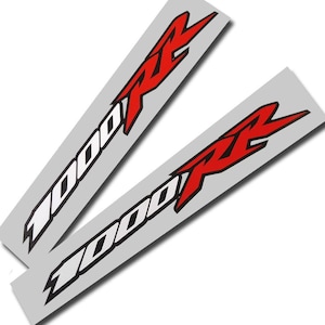 Op de afbeelding: Twee zilverkleurige stickers met de tekst "1000 RR" in een vetgedrukte, gestileerde lettertype. De "1000" is wit met een zwarte omlijning en de "RR" is rood met een zwarte omlijning. De stickers zijn schuin geplaatst en overlappen elkaar licht.