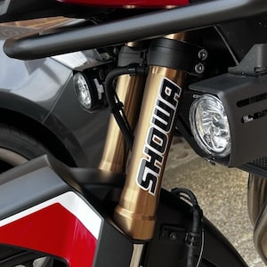 Peut inclure: Gros plan sur la suspension avant d'une moto. Les tubes de fourche dorés portent le nom de marque "SHOWA" en blanc. La moto a un garde-boue noir avec des accents rouges et blancs, et un guidon noir. Deux phares sont visibles.