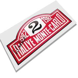 May include: A rectangular sticker with a red and white design. The sticker features the text "RALLYE MONTE-CARLO" in white, with the number "2" in black and the year "1982". A signature is visible in the lower left corner.