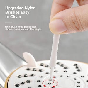 Op de afbeelding: Een witte reinigingsborstel met nylon haren die op een douchekop wordt gebruikt. De tekst luidt: Upgraded Nylon Bristles Easy to Clean. Fine brush head penetrates shower holes to clean blockages. Het hulpmiddel heeft een slanke steel.