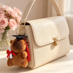 May include: Cream-colored handbag with a gold clasp and handle. A graduation-themed teddy bear keychain with a black cap, red tassel, and diploma hangs from the bag. A bouquet of pink roses is in the background.
