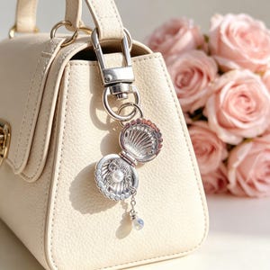 May include: A silver seashell charm with a pearl and a dangling iridescent bead is attached to a cream-coloured handbag. The charm is open, revealing the pearl inside. A bouquet of pink roses is in the background.