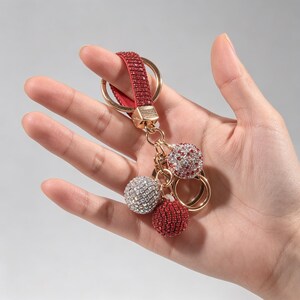 May include: A keychain featuring a red strap encrusted with small red crystals, a gold-tone metal ring, and three spherical charms. One charm is covered in clear crystals, one in red crystals, and one in a mix of red and clear crystals.
