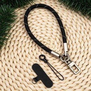 May include: A black rope wrist strap featuring a reflective white thread pattern, connected to a silver-toned metal swivel lobster clasp. Included is a small black cord loop attachment and a black rectangular tab with a metal ring for secure fastening.