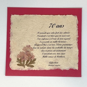 Può includere: Un biglietto di auguri fatto a mano con un bordo rosso e un centro strutturato bianco sporco. Il biglietto presenta fiori secchi e testo francese che celebra un 70° compleanno. Il testo include le parole "70 ans" e una poesia.