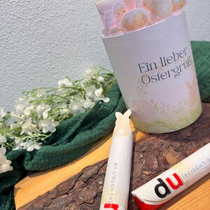 May include: A white and pink Easter-themed gift container with the text "Ein lieber Ostergruß". The container is filled with Easter eggs and bunny cutouts. Two white and red candy sticks with the word "du" are also visible.