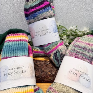 Könnte beinhalten: Handgestrickte Cozy Socken in verschiedenen Farben. Die Socken haben Streifen in Pink, Blau, Gelb und Grün. Jedes Paar ist mit einem Etikett mit der Aufschrift "Hand Knitted Cozy Socks" versehen. Die Socken sind aus weichem, warmem Material.