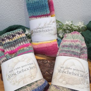 May include: Three pairs of colorful, hand-knitted socks with the text "mit Liebe gestrickte Kuschelsocken" on the labels. The socks feature various color combinations, including blue, pink, yellow, and green. The socks are displayed on a wooden surface.