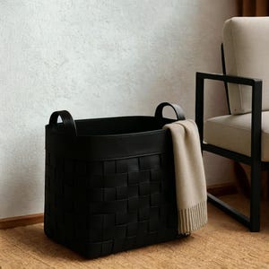 Może przedstawiać: Czarny, tkany kosz do przechowywania z dwoma uchwytami. Kosz ma prostokątny kształt i jest wykonany z ciemnego materiału. Beżowy koc jest zarzucony na bok kosza. Kosz stoi na tkanej macie.