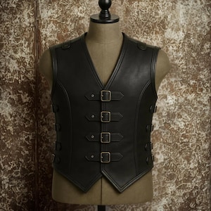 Puede incluir: Chaleco de cuero negro con cuello en V y hebillas color latón. El chaleco tiene un corte entallado y costuras decorativas. El chaleco tiene tres hebillas en la parte delantera y dos a cada lado. El chaleco se exhibe en un maniquí.