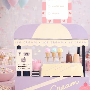 Könnte beinhalten: Ein dekorativer Eisstand in Lila und Creme. Der Stand hat eine Markise, ein "Ice Cream"-Schild und ein Bestellformular. Drei Eiswaffeln werden zusammen mit Toppings ausgestellt. Ballons und andere Leckereien sind im Hintergrund.