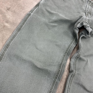 May include: Close-up of sage green denim pants. The pants have visible stitching along the seams and pockets. The fabric appears to be slightly worn, with subtle variations in color. The pants are folded, showing the texture and details of the denim.