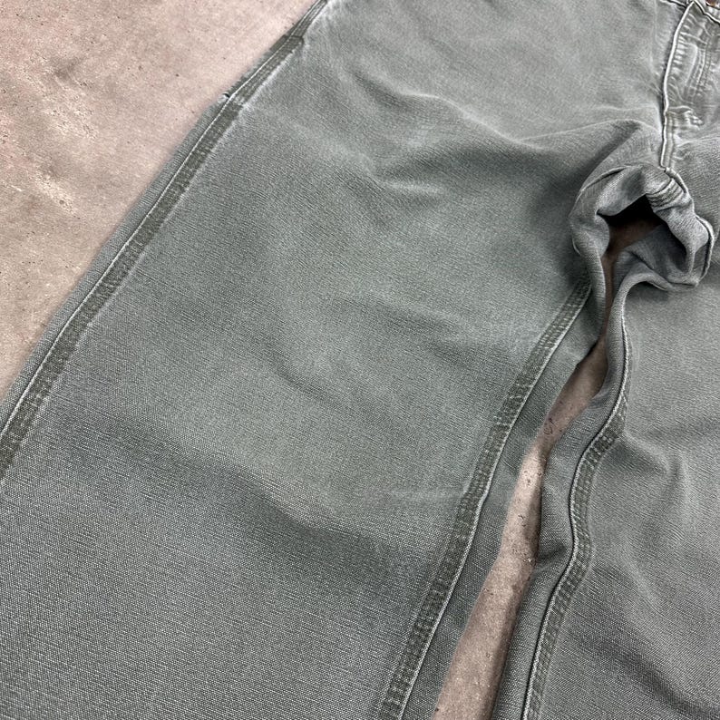 May include: Close-up of sage green denim pants. The pants have visible stitching along the seams and pockets. The fabric appears to be slightly worn, with subtle variations in color. The pants are folded, showing the texture and details of the denim.