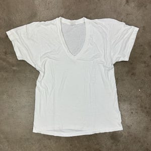 May include: A white V-neck t-shirt with short sleeves. The shirt is made of a soft, lightweight fabric and is laid flat on a neutral surface. The neckline is a deep V-shape.