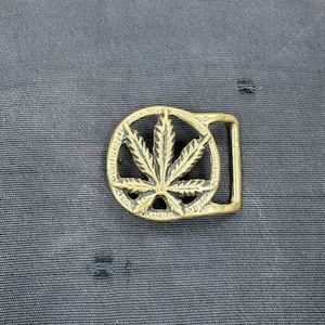 May include: A brass belt buckle with a cannabis leaf design inside a circular frame. The buckle has a rectangular bar for the belt strap. The background is a dark, textured fabric.