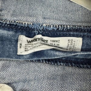 K&ouml;nnte beinhalten: Nahaufnahme eines blauen Jeanskleidungsst&uuml;cks mit einem wei&szlig;en Etikett. Das Etikett lautet "Wrangler 13MWZ 33 X 38" und "Made in Costa Rica". Der Denim hat ein strukturiertes Aussehen.