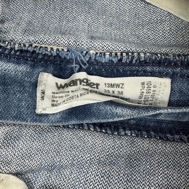 K&ouml;nnte beinhalten: Nahaufnahme eines blauen Jeanskleidungsst&uuml;cks mit einem wei&szlig;en Etikett. Das Etikett lautet "Wrangler 13MWZ 33 X 38" und "Made in Costa Rica". Der Denim hat ein strukturiertes Aussehen.